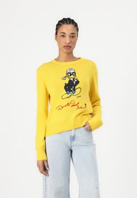 Pull en tricot jaune avec ourlets et poignets côtelés, orné d'un graphique coloré de Donald Duck et de sa signature brodée sur le devant.