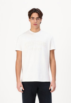 Hvid bomulds t-shirt med "TOMMY JEANS New York" logo i beige broderi, rund hals og korte ærmer, parret med mørke jeans.