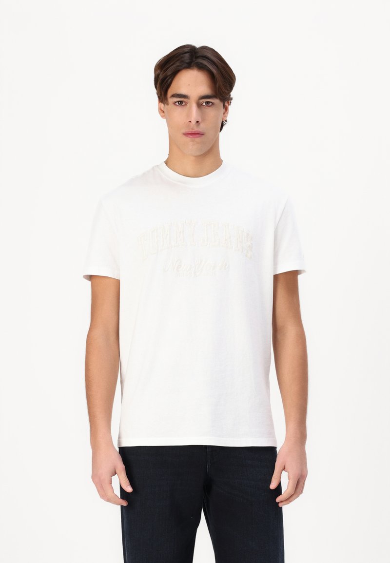 Witte katoenen T-shirt met het "TOMMY JEANS New York" logo in beige borduursel, ronde hals, en korte mouwen, gedragen met donkere jeans.