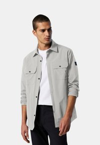 Camicia grigia chiaro con maniche lunghe, due tasche frontali e dettagli neri sui bottoni. Indossata sopra una maglietta bianca, abbinata a pantaloni scuri.