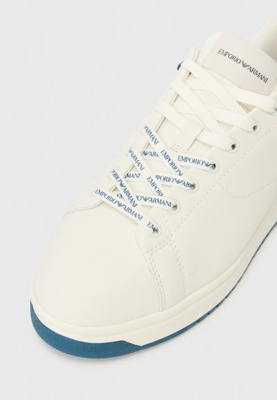 Vita lädersneakers med blå mocka sula. Har blåa tryckta snörningar och en tagg präglad med varumärket. Rundad tådesign.
