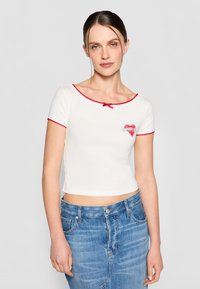Hvit kort topp med korte ermer, rød bølget kant og en sentral hjertegrafikk med "Tommy." Brukt sammen med høyt midje blå denimskjørt.