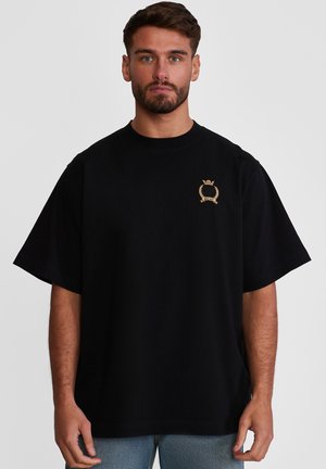 Mann mit kurzen Haaren und Bart, trägt ein schwarzes, übergroßes T-Shirt mit kleinem goldenem Emblem auf der linken Brust, blaue Jeans, neutraler Hintergrund.