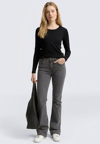 Zwart top met lange mouwen gecombineerd met grijze uitlopende jeans, gedragen met witte sneakers en een zwarte schoudertas vasthoudend, tegen een lichte achtergrond.