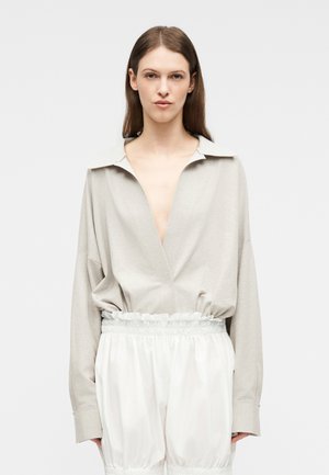 Ženska, oblečena v ohlapno bež bluzo z V-izrezom in širokim ovratnikom ter bele visoke hlače z elastičnim nabranim pasom.