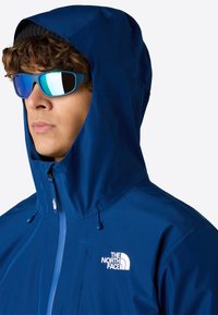 Osoba v modré bundě s kapucí od značky North Face a reflektivních modrých slunečních brýlích, obrácená vlevo, s částečně viditelnými kudrnatými hnědými vlasy.