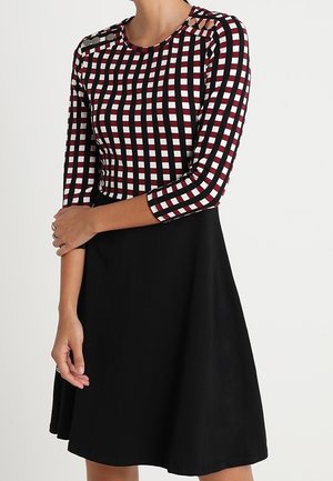 Kobieta ubrana w sukienkę z czarną spódnicą oraz czerwoną, białą i czarną kratkowaną górą z rękawami do łokcia i guzikami na ramionach.