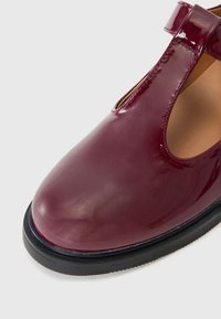 Chaussure à bride en T rouge foncé brillante avec bout arrondi et semelle noire, gros plan sur la partie avant supérieure et le détail de la boucle de la bride.
