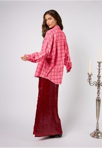Różowa, oversize'owa koszula w kratę z długimi rękawami, zestawiona z długą czerwoną spódnicą z cekinami. Koszula posiada kołnierzyk zapinany na guziki oraz luźny krój.