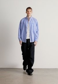 Gestreiftes blaues Hemd mit Knöpfen, lockerer Passform, kombiniert mit dunklen Jeans und schwarzen Schuhen. Das Hemd verfügt über eine Tasche und weiße Knöpfe.