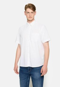 Camisa blanca de manga corta con botón y cuello, que presenta un bolsillo en el pecho y una tela ligera y texturizada. Lleva puesta unos pantalones vaqueros azules.
