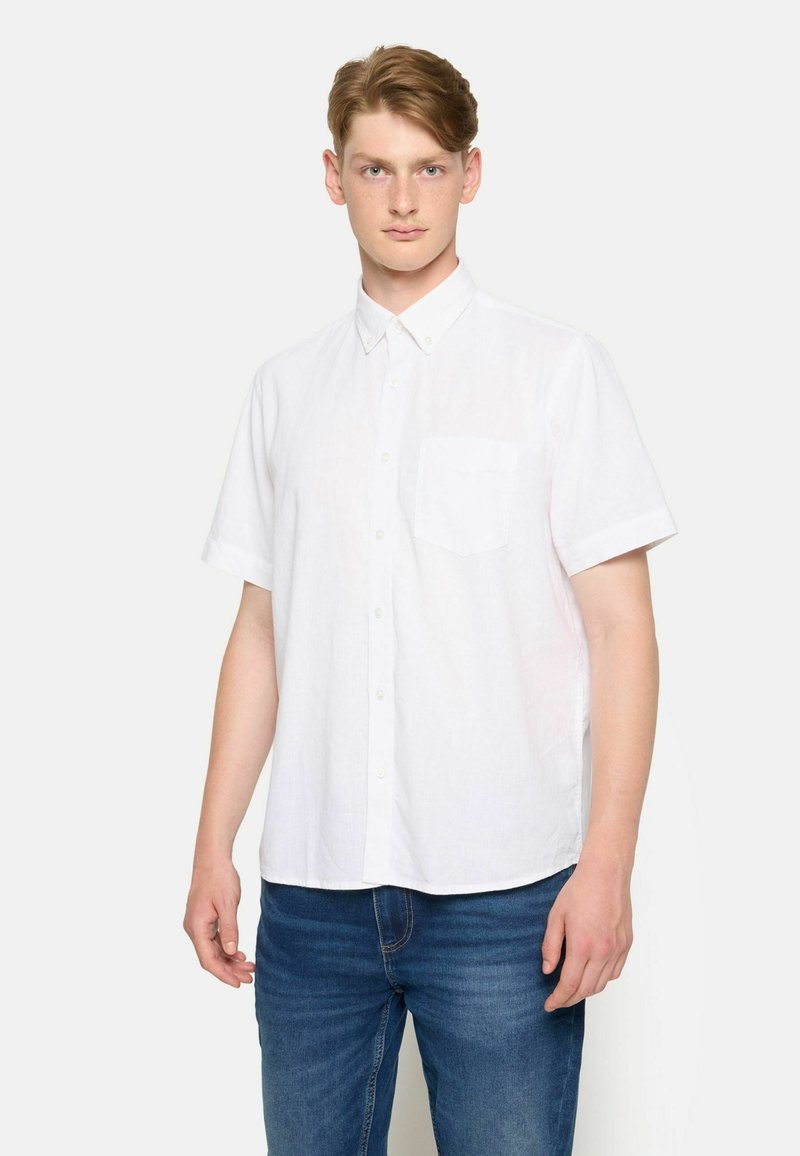 Camisa blanca de manga corta con botón y cuello, que presenta un bolsillo en el pecho y una tela ligera y texturizada. Lleva puesta unos pantalones vaqueros azules.