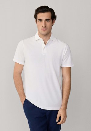 Jonge man staat met één hand in zijn zak, gekleed in een wit polohemd met korte mouwen en donkerblauwe broek, tegen een effen achtergrond.