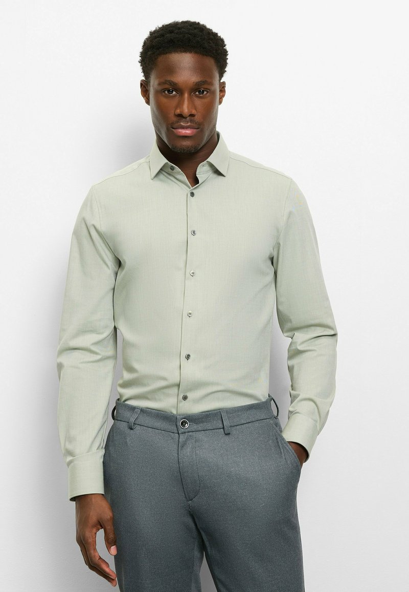 Hellgrünes Hemd mit langen Ärmeln und Button-Down-Kragen, aus strukturiertem Stoff mit grauen Knöpfen, kombiniert mit grauer Hose.