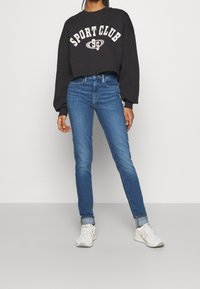 Sudadera negra corta con estampado "SPORT CLUB", combinada con jeans ajustados azules. Modelo lleva zapatillas blancas. Conjunto sencillo y casual.