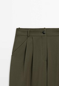 Pantalon vert olive sur mesure avec plis, passants de ceinture, poche latérale avant et fermeture à bouton noir unique à la taille.