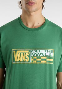Camiseta de algodón verde con un gran logo de Vans en amarillo y blanco, con el texto "OFF THE WALL" y un patrón a cuadros en la parte inferior.