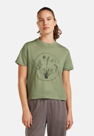 Young woman with brown hair in ponytail wearing a green t-shirt with botanical sketches and gray pants, standing against a plain background.