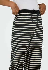 Black and cream striped pants with a soft texture, elastic waistband, and adjustable drawstring. Sides feature pockets for functionality.