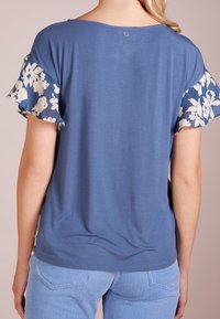Top azul con mangas de estampado floral. Tejido suave, corte holgado, escote redondeado con sutil detalle en la parte posterior. Textura ligera y transpirable.