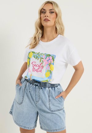 Vrouw draagt een wit T-shirt met de tekst "Sicily Italia" en een design van citroenbloemen, gecombineerd met lichtblauwe hooggetailleerde spijkershorts.