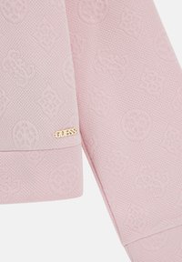 Tessuto di colore rosa chiaro caratterizzato da motivi goffrati e un dettaglio del logo "GUESS" in oro su un orlo piegato.