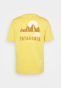 Camiseta de algodón amarilla con un gráfico de montaña marrón, un sol, el texto "PATAGONIA" y "Desde 1973" debajo. Mangas cortas, cuello redondo.