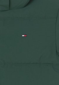 Tessuto verde scuro con piccolo logo ricamato rettangolare in rosso, bianco e blu al centro, con linee di cucitura orizzontali sopra e sotto.