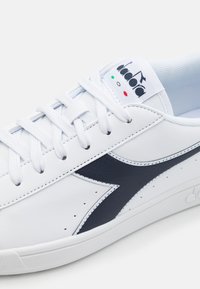 White leather sneaker with navy accent, round toe, perforations, flat laces, and logo on tongue. Smooth texture and low-top design.