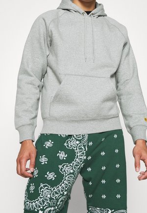 Person trägt einen hellgrauen Kapuzenpullover mit Fronttasche und dunkelgrüne Hose mit weißen Paisley- und geometrischen Mustern.