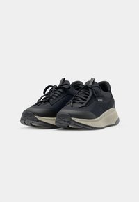 Black sports shoes with textured synthetic upper, round toe, laces, and a chunky beige sole. Features brand logo on the side.