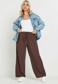 Pantalones anchos marrones con cintura elástica, combinados con una camiseta sin mangas blanca y una chaqueta de mezclilla azul claro. Zapatillas blancas completan el conjunto.