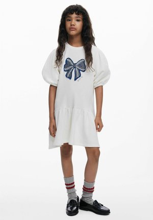 Biele šaty s naberanými rukávmi a modrým vyšívaným detailom v tvare mašle, rozšíreným lemom, v kombinácii so sivými ponožkami s červenými pruhmi a čiernymi mokasínami.