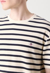 Striped t-shirt in cream and navy, featuring a ribbed texture, round neckline, and a logo on the chest. Short sleeves, casual fit.
