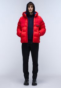 Röd pufferjacka med huva, med en strukturerad yta och en logotypapplikation. Bärs över en marinblå hoodie och svarta byxor, med svarta skor.