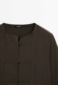Chemisier marron foncé avec un col rond, doté d'une patte de boutonnage à l'avant, de trois boutons et d'une finition texturée subtile.