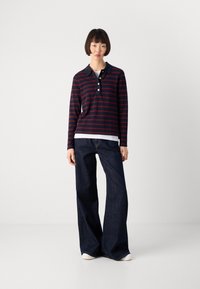 Woman wearing a navy and burgundy striped long-sleeve polo shirt, dark wide-leg jeans, and white sneakers standing against a white background.