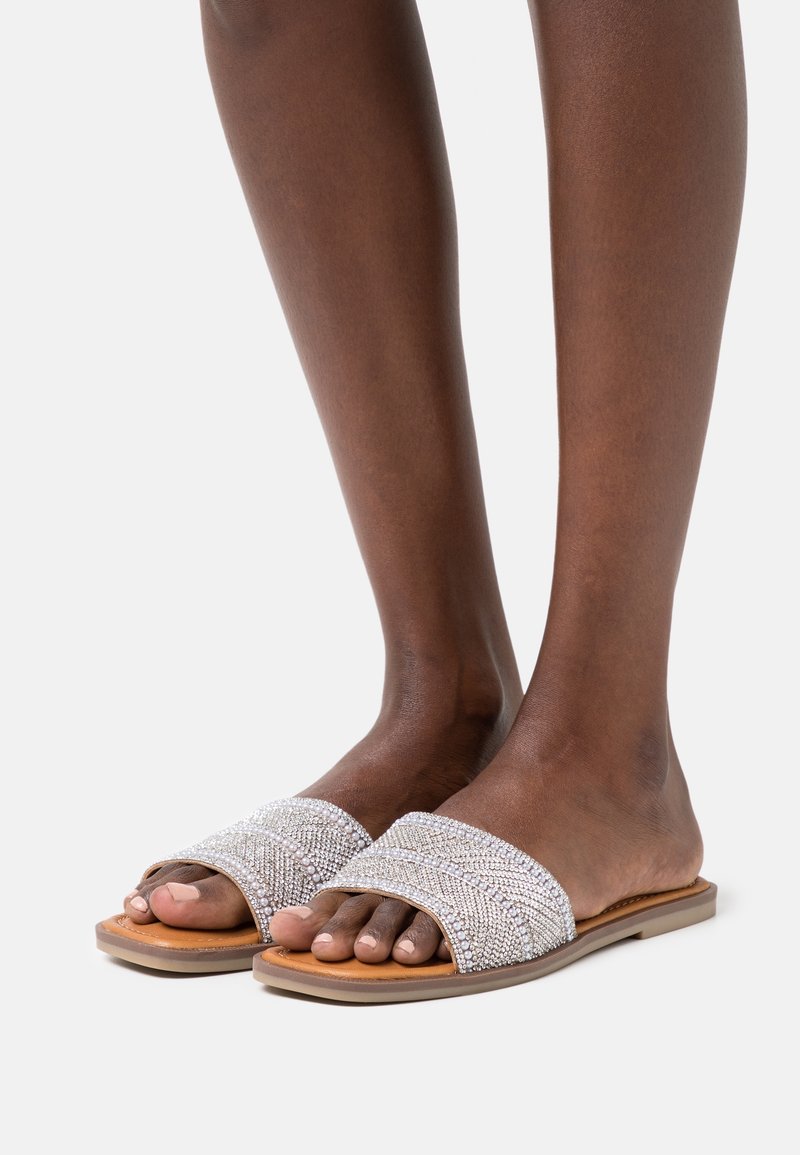 Pieds portant des sandales slides couleur beige avec de larges brides recouvertes de strass argentés sur fond blanc.