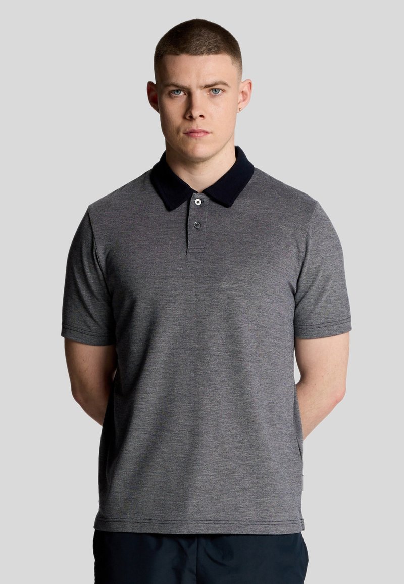 Grijze poloshirt van textuurstof, met een marineblauwe kraag, drie knopen en korte mouwen. Model staat recht voor zich uit.