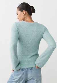 Mintgrüner, flauschiger Pullover mit langen Ärmeln, gerippten Bündchen und rundem Ausschnitt. Weiche Textur mit entspannter Passform. Kombiniert mit hellblauen Jeans.
