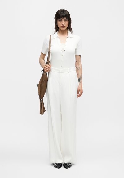 Γυναίκα με κοντά σκουρόχρωμα μαλλιά, φοράει λευκό κοντομάνικο πουκάμισο με κουμπιά και φαρδιά λευκά παντελόνια, κρατώντας καφέ τσάντα ώμου με φούντες.