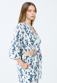 Weißes Oberteil aus Stoff mit blauen Blumenmustern, tiefer V-Ausschnitt, kurze Ärmel, geknotetes Vorderdetail, kombiniert mit passenden gemusterten Shorts.