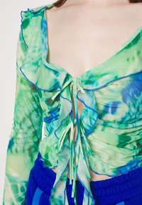 Green and blue sheer blouse with ruffled edges, tie closure at the front, and long sleeves. Textured, lightweight fabric with a floral pattern.