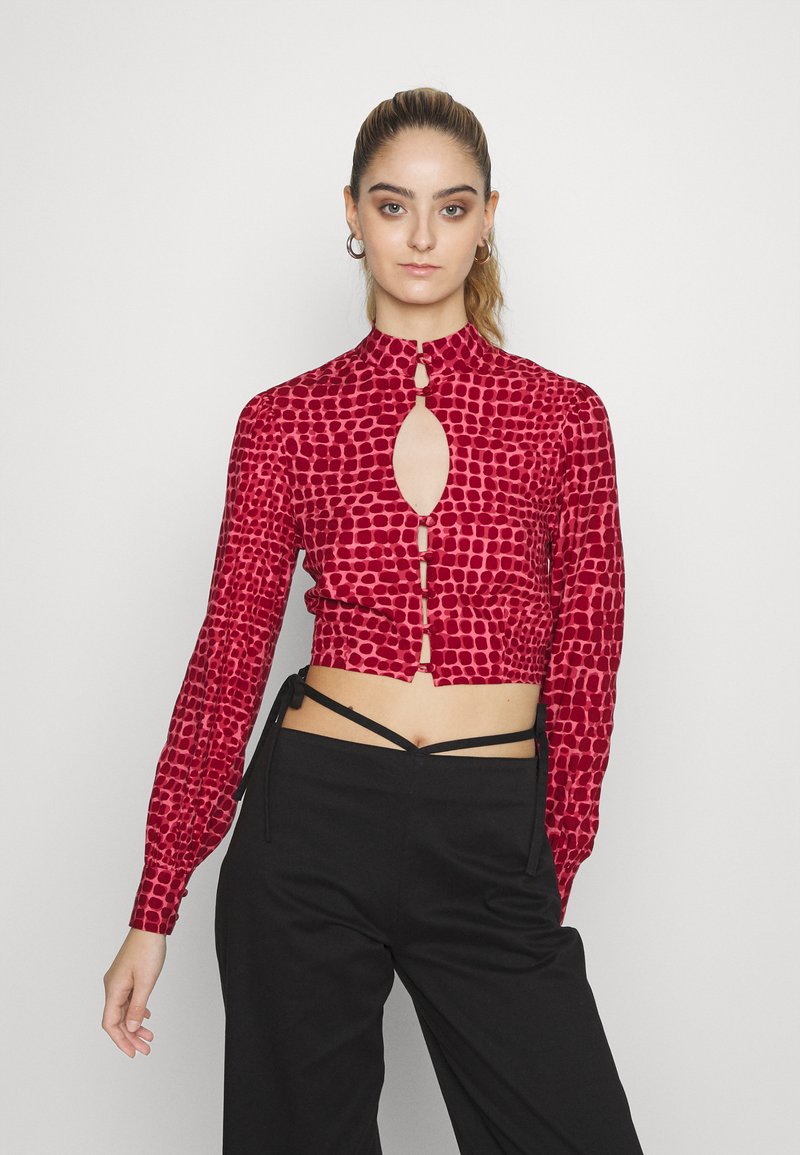 Blusa curta vermelha com um padrão animal exclusivo, gola alta, recorte em forma de buraco de chave e detalhes em botão na parte da frente. Mangas longas e ajustadas.
