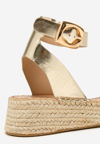 Sandalia de espadrille metálica dorada con una amplia correa para el tobillo, que presenta una hebilla cuadrada y una suela de plataforma de yute tejido. Textura suave.
