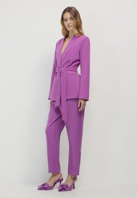 Donna che indossa un blazer viola con cintura e pantaloni abbinati, insieme a scarpe con tacco a stiletto viola, in piedi contro uno sfondo bianco uniforme.