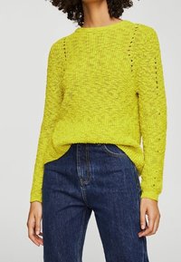 Limonengrüner Strickpullover mit rundem Halsauschnitt, der an den Schultern mit offenem Strickmuster versehen ist, kombiniert mit dunkelblauen hochgeschnittenen Jeans.