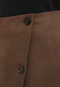 Καφέ σουέντ ύφασμα με απαλή υφή, που διαθέτει μαύρο κουμπί στο κλείσιμο και καθαρό, ραμμένο σχέδιο κατά μήκος της άκρης.