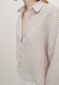 Personne portant une chemise beige et blanche à rayures verticales avec des boutons et un col ouvert, visible des épaules à la taille sur un fond uni.