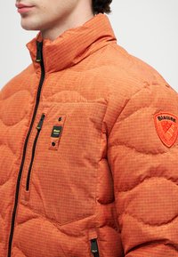 Narančasta puffa jakna s kariranim uzorkom, s visokim ovratnikom, zatvaranjem na patentni zatvarač i logom na patchu na ramenu.